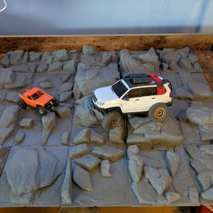 May include: An overhead shot of a miniature off-road terrain with two toy vehicles. One is an orange buggy, and the other is a white SUV. The terrain is made of gray, rock-like tiles.