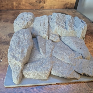 May include: A gray, 3D-printed miniature rock formation with a textured surface. The model features multiple rock shapes and sizes, suitable for tabletop gaming or model scenery.