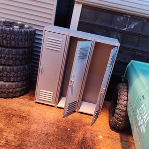 May include: A set of three grey metal lockers with one door open. The lockers are on a wooden surface with a stack of tyres and a toy truck in the background.