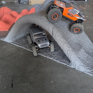 Può includere: Una rampa per percorsi a ostacoli grigia con una texture simile alla roccia, con un veicolo fuoristrada radiocomandato grigio posizionato sotto e un camion radiocomandato arancione e nero che sale sulla pendenza esterna. Un pezzo di terreno arancione si trova sullo sfondo.
