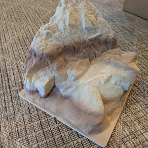 May include: A detailed, textured mountain model in shades of beige, brown, and cream, mounted on a square, off-white base. The miniature landscape features rugged peaks and valleys, perfect for display or educational purposes.