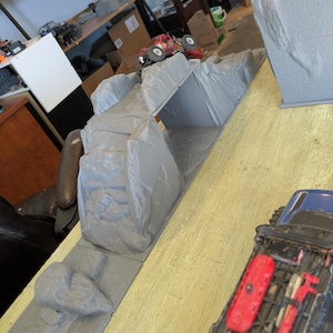 May include: A grey 3D printed rock crawling obstacle course featuring a bridge section and textured terrain. A small red remote-controlled vehicle is positioned on the elevated rock formation, while a blue vehicle is visible in the foreground.