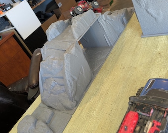 May include: A grey 3D printed rock crawling obstacle course featuring a bridge section and textured terrain. A small red remote-controlled vehicle is positioned on the elevated rock formation, while a blue vehicle is visible in the foreground.