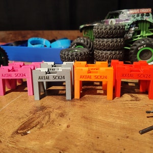 May include: Five 3D printed stands for a toy car. The stands are in different colors: pink, gray, orange, and red. Each stand has the text "AXIAL SCX24" printed on it.