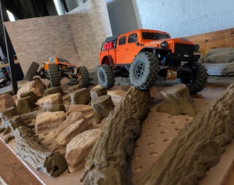 Könnte beinhalten: Zwei orangefarbene ferngesteuerte Trucks fahren über ein Miniatur-Gelände aus Felsen und Baumstämmen. Die Trucks haben große, grobstollige Reifen, und der Vordergrund zeigt detaillierte Miniatur-Baumstämme und Felsen. Die Szene spielt vor dem Hintergrund einer Backsteinmauer.