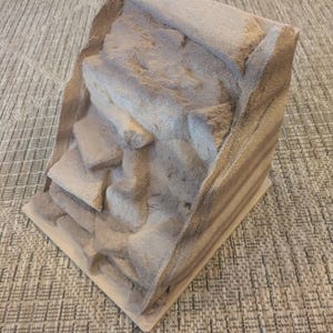 以下が含まれることがあります： 層状で階段状のデザインが施された、ミニチュアのテクスチャード加工された風景モデル。モデルは、岩の多い地形に似た、さまざまなベージュと茶色の色合いが特徴です。ベースは明るい色の正方形で、全体的な構造はジオラマまたはディスプレイピースを示唆しています。