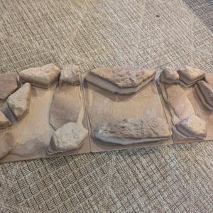 Puede incluir: Un fondo decorativo para terrario con un diseño de rocas y piedras. El fondo está hecho de múltiples paneles y presenta una variedad de formaciones rocosas en tonos beige y gris. El tema general es natural y terroso.