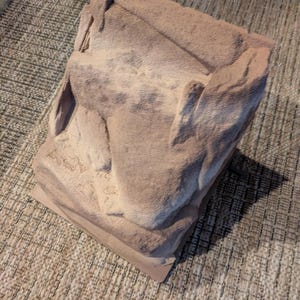 Puede incluir: Una escultura rectangular texturizada, de color marrón claro, con un aspecto rugoso y tallado. La escultura tiene un diseño en capas y parece estar hecha de piedra o un material similar. La pieza está sobre una alfombra tejida de tonos neutros.