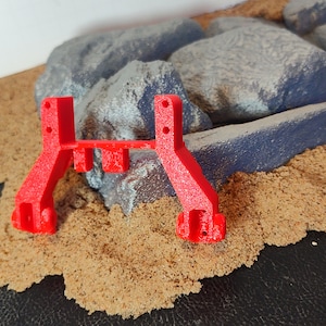 以下が含まれることがあります： 2本の垂直な支柱と水平な接続部を持つ、赤色のプラスチック製の3Dプリント部品。部品は茶色の表面の上に置かれています