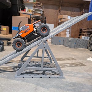 May include: An orange and black remote-controlled rock crawler vehicle with the text XC-1 on the side, positioned on a grey plastic adjustable ramp structure. The vehicle features large, textured black tyres with orange rims and a roll cage design.