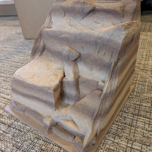 May include: A miniature rock formation model in shades of brown and beige. The textured surface has a layered appearance, with a flat base and a rugged, uneven surface, resembling a natural landscape feature.