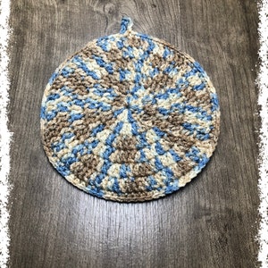 May include: A round, crocheted hot pad in shades of blue, beige, and brown. The circular design features a loop for hanging. The handmade item is suitable for kitchen use and adds a touch of rustic charm.
