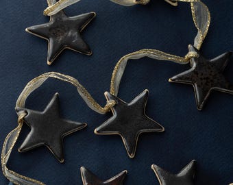 May include: A collection of dark ceramic star ornaments strung on a sheer ribbon with gold accents. The stars have a glossy finish and are arranged against a dark blue background, creating a festive and decorative display.