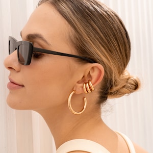 May include: A woman wearing black sunglasses and gold hoop earrings. The earrings are stacked in a graduated size, with the largest hoop being the bottom earring.