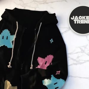 Puede incluir: Sudadera negra con cremallera y diseños de estrellas pixeladas de colores. La sudadera con capucha tiene una capucha con cordón y cierre de cremallera. El diseño incluye estrellas celeste, rosas y amarillas. El logo "JACKET TRENDY" está en un círculo negro.