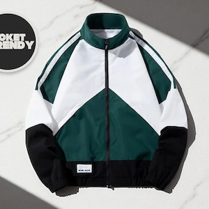 Può includere: Giacca sportiva con collo alto e cerniera a tutta lunghezza. La giacca presenta un design a blocchi di colore con pannelli bianchi, verde scuro e neri. Il testo "JACKET TRENDY" è in un cerchio nero in alto a sinistra.