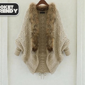 May include: A beige knitted cardigan with a faux fur collar, hanging on a wooden hanger. The cardigan has a cable knit pattern and a drawstring closure. The text "JACKET TRENDY" is in a black and white rectangle in the upper left corner.