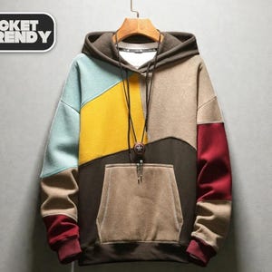 Puede incluir: Una sudadera con capucha de bloques de color con capucha marrón, un panel azul claro y amarillo y un bolsillo marrón. Las mangas son marrones y rojas. El texto "JACKET TRENDY" está en un cuadro blanco.