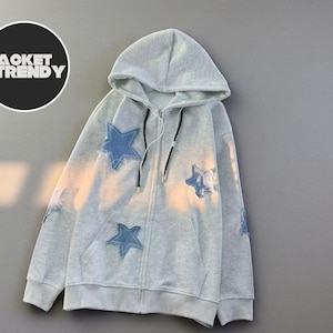 Puede incluir: Una sudadera con capucha gris claro con cremallera, capucha y cordones negros. La sudadera con capucha presenta parches de estrellas azules en la parte delantera y en las mangas. El texto "JACKET TRENDY" está en un círculo negro.