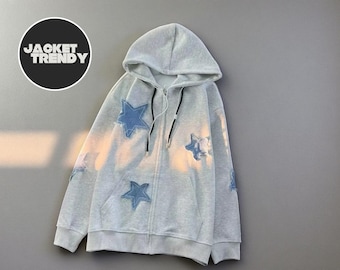 May include: A light gray zip-up hoodie with a hood and black drawstrings. The hoodie features blue star patches on the front and sleeves. The text "JACKET TRENDY" is in a black circle.