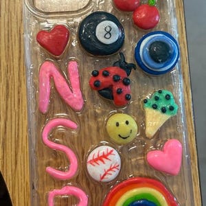 May include: Clear phone case decorated with various colorful charms. Includes a rainbow, a baseball, a smiley face, a ladybug, a heart, a cherry, a number 8 ball, and the letters NSP.