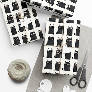 May include: Wrapping paper featuring a repeating pattern of black cats with orange noses on a white background. The paper is tied with brown twine and includes white star and heart-shaped gift tags, a ball of yarn, and metal scissors.