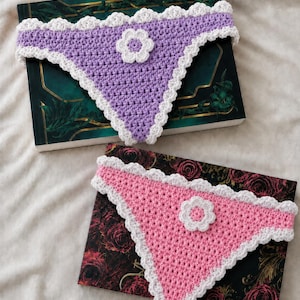 May include: Two triangular crochet bandanas, one in lavender and one in pink, each featuring a white scalloped border and a central white flower appliqué. The lavender bandana is placed on a dark green book, and the pink one on a floral book.