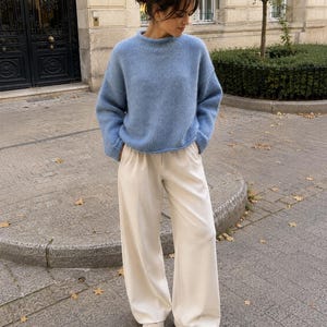 May include: A light blue long-sleeved knit sweater with a crew neckline paired with high-waisted cream-colored wide-leg trousers and white sneakers. The sweater has a soft, fuzzy texture and the trousers feature a clean, tailored silhouette.