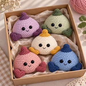 Puede incluir: Cinco pequeños juguetes de peluche tejidos a ganchillo, redondos, con puntas y pies en forma de corazón, dispuestos en una caja de cartón. Los juguetes son morados, verde claro, crema, rosas y azules, con dos ojos negros y una sonrisa.