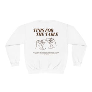 May include: White sweatshirt with brown text that reads "TINIS FOR THE TABLE" and a line drawing of two hands holding up martini glasses.