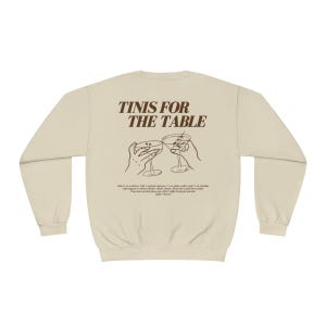 May include: A beige sweatshirt with the text "TINIS FOR THE TABLE" printed in brown on the back. The text is surrounded by a graphic of two hands holding up martini glasses.