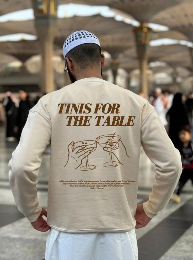 May include: A beige sweatshirt with brown text that reads "TINIS FOR THE TABLE" and a graphic of two hands holding up martini glasses. The text below the graphic reads "Add ice to a shaker, add 1 oz fresh expresso, 1 oz plain vanilla and 1 oz. Kahlua (give a vigorous shake to shaker shake, shake, shake, shake for a good 30 seconds. Pour two martini glasses with 2 coffee beans for garnish. Enjoy! Cheers!"