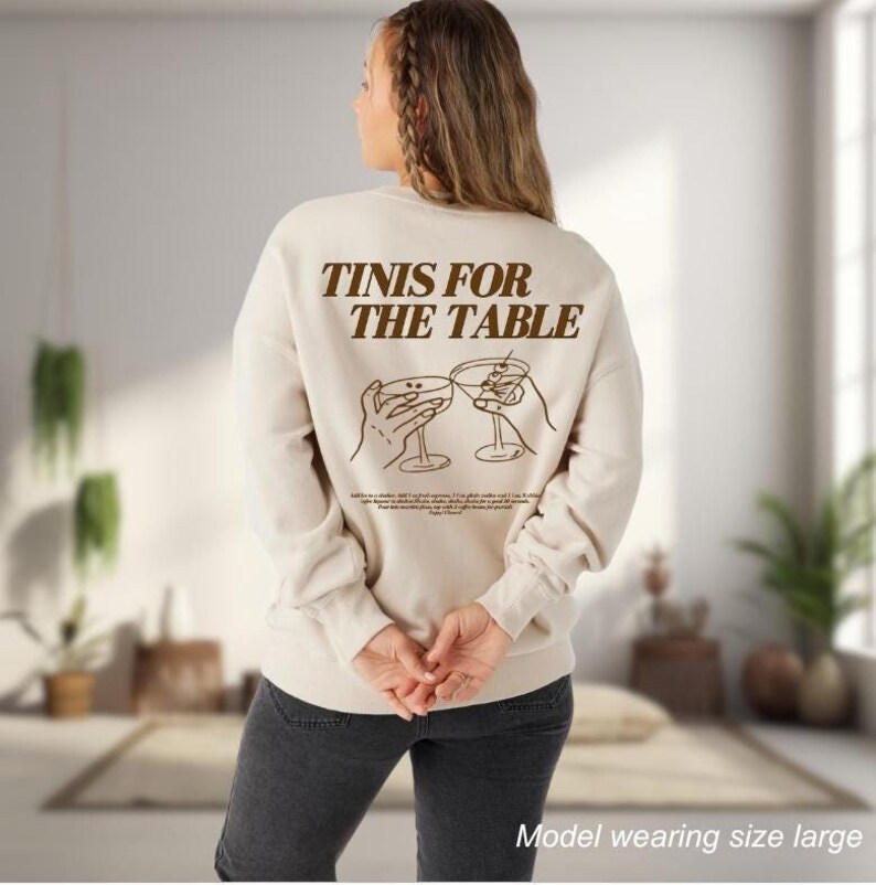 May include: A cream colored sweatshirt with brown text that reads "TINIS FOR THE TABLE" and a graphic of two hands holding up martini glasses. The model is wearing a size large.