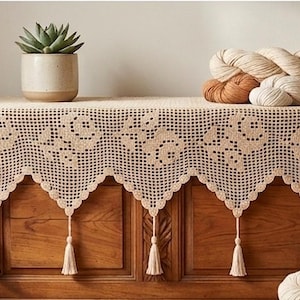 May include: A wooden chest is topped with a cream-colored, crocheted table runner with a scalloped edge and tassels. A potted succulent and several skeins of yarn in neutral tones sit on top. The chest is made of wood.