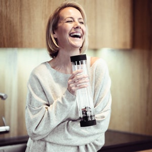 Könnte beinhalten: Eine Frau in einem hellgrauen Pullover lacht, während sie eine durchsichtige Glasflasche mit schwarzem Deckel und einem schwarz-weißen Design im Inneren hält. Die Flasche ist etwa 25 cm hoch.