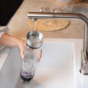 Könnte beinhalten: Eine klare Glaswasserflasche mit einem Edelstahldeckel und einem geometrischen Muster im Inneren. Wasser wird aus einem silbernen Wasserhahn in die Flasche gegossen.