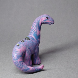 May include: A small dinosaur figurine featuring a marbled pattern of pink and blue swirls with embedded glitter. The creature has a long neck, a curved tail, and three small, dark, reflective circular accents along its back and spine.