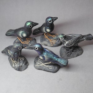May include: Six decorative bird figurines with iridescent blue and green feathers, perched on gray stone bases. The birds have white accents and wire legs. The figurines are arranged on a gray surface.