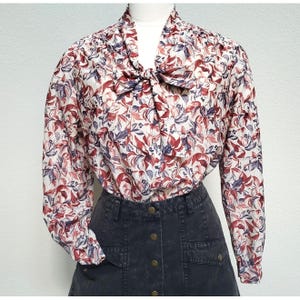 May include: A long-sleeved blouse with a floral pattern in red, burgundy, and blue on a cream background. The blouse features a bow at the neckline and is paired with a dark denim skirt. The blouse has a vintage aesthetic.