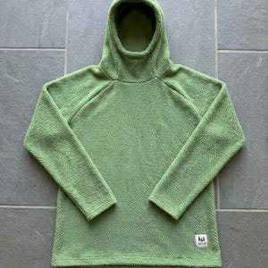 May include: A light green long-sleeve hooded pullover made of a textured fleece material. The garment features a hood, raglan sleeves, and a small rectangular white patch on the lower front corner with a black graphic of three trees and text.