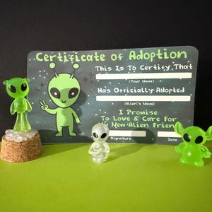 May include: Three small alien figurines and a rectangular Certificate of Adoption card. One green alien stands on a cork base, one clear alien stands in the center, and one green alien stands on the right. The card features alien graphics and text.