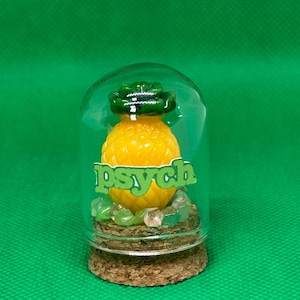 May include: A glass jar with a cork lid contains a yellow pineapple figurine with green leaves. Green and pink stones are at the base of the pineapple. The word "psych" is written on the jar in green letters.
