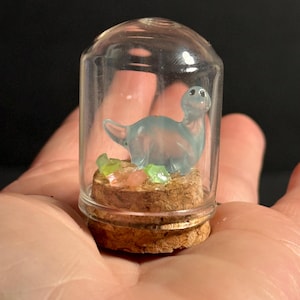 May include: A small glass jar with a cork lid contains a light blue glass dinosaur figurine. The dinosaur is standing on a bed of green and pink glitter.