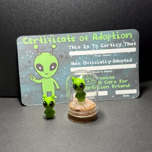 May include: A small green alien figurine stands next to a glass dome containing a second green alien figurine on a cork base. Behind them is a rectangular card titled Certificate of Adoption with a green alien illustration and blank lines.