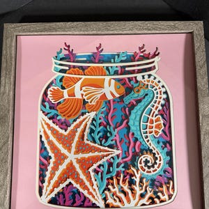 May include: A framed paper art piece depicting a glass jar filled with colorful sea life. The jar contains a starfish, a seahorse, and a clownfish, all surrounded by coral and seaweed.
