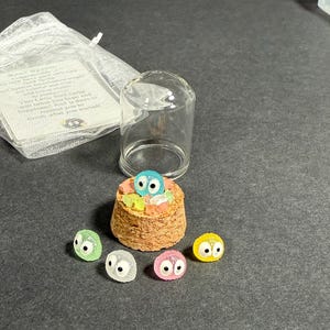 May include: Five small, colorful, glass figurines with large eyes, resembling the characters from the movie 'My Neighbor Totoro'. The figurines are sitting on a cork base with a glass dome over them.