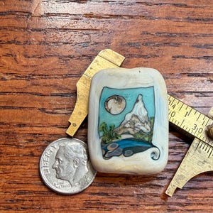 May include: A rectangular off-white bead featuring a miniature landscape scene with a mountain, a moon, and a blue body of water. The bead is positioned next to a US dime and a brass caliper, which shows the bead is approximately 0.75 inches wide.