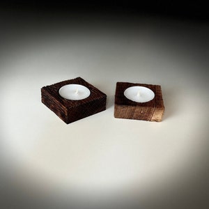 May include: Two wooden candle holders with a square base and a round recess for a tea light candle. The holders are made of dark brown wood and are sitting on a white surface.