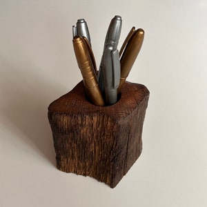 Puede incluir: Un portaplumas de madera hecho de un tronco de árbol con un acabado natural de corteza. El portaplumas está lleno de bolígrafos dorados y plateados.