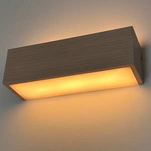 Puede incluir: Aplique de pared rectangular de madera clara que emite una luz cálida y difusa. La luminaria está fijada a una pared blanca. La fuente de luz es un panel largo y rectangular que emite un brillo suave y acogedor.